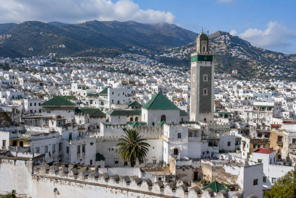 Panoramic view of a white Moroccan town with green-tiled roofs, a tall minaret-like tower, and surrounding walls against a mountain backdrop